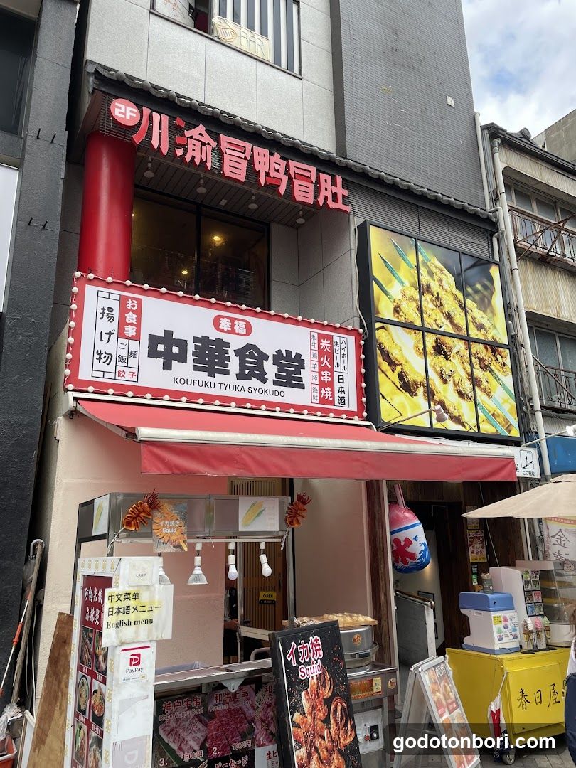 A street-level view of a Japanese konbini-style shop named 'Koufuku Tyuka Syokudo' with signage in Chinese and Japanese characters. The storefront features red awnings, illuminated signs displaying the name and menu items like 'Yaki Onigiri', and large windows showing food inside. A yellow cart is visible to the right.