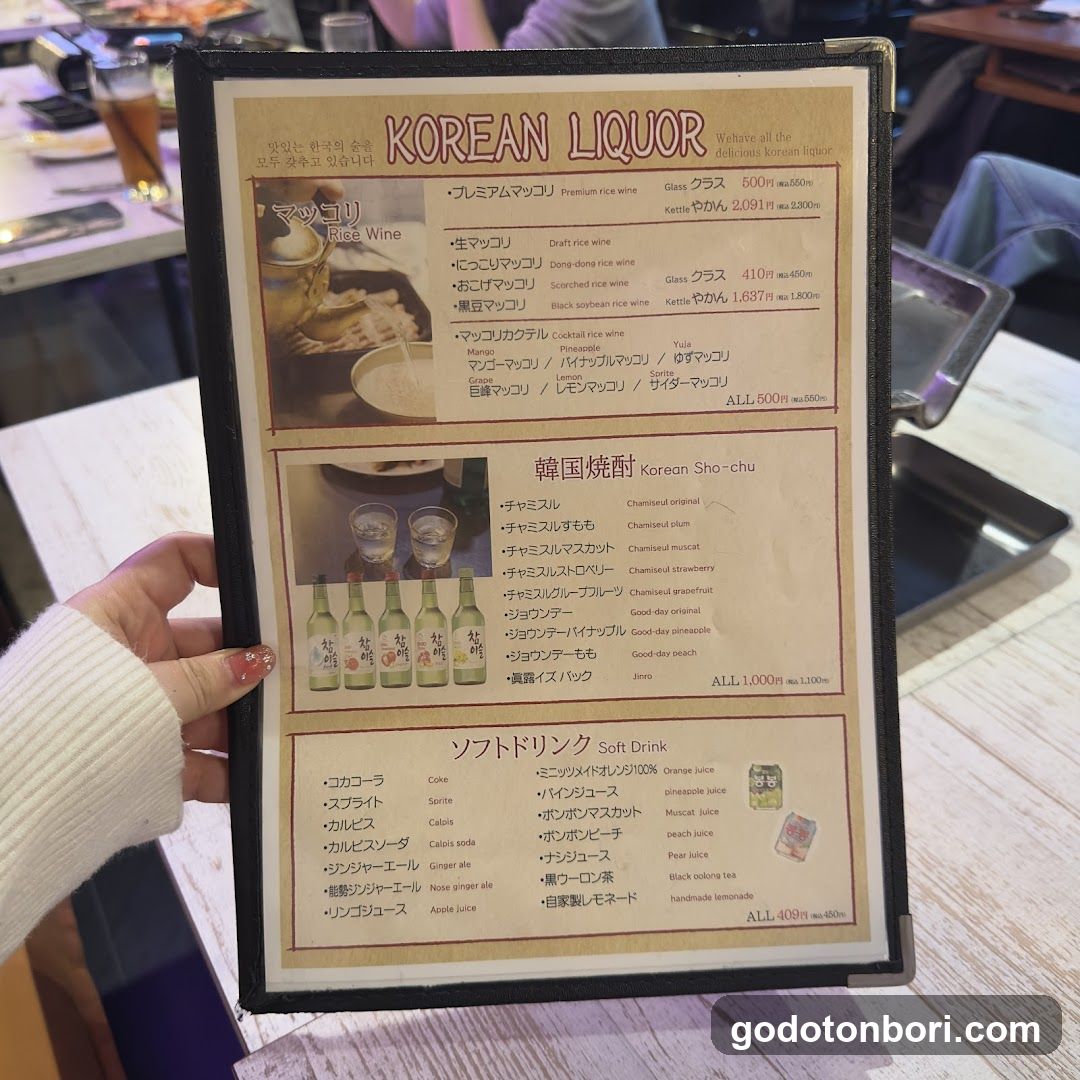A close-up photo of a framed menu board displaying Korean liquor and soft drink options, held by a person in a white sweater.
