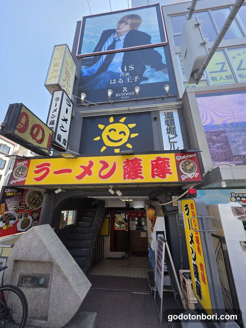 A street-level view of a ramen shop entrance with bright yellow signage reading 'ラーメン藤原' (Ramen Furuma). Above the sign, there is a large billboard featuring an idol group and Japanese text. A smiling sun logo is visible on the building facade. To the left, a stone monument with Japanese characters stands near a bicycle wheel. Stairs lead up to the shop entrance.