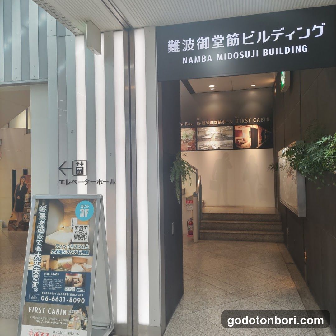 Entrance to Namba Midosuji Building with visible signage and elevator hall sign. A promotional standee for First Cabin hotel is placed near the entrance.