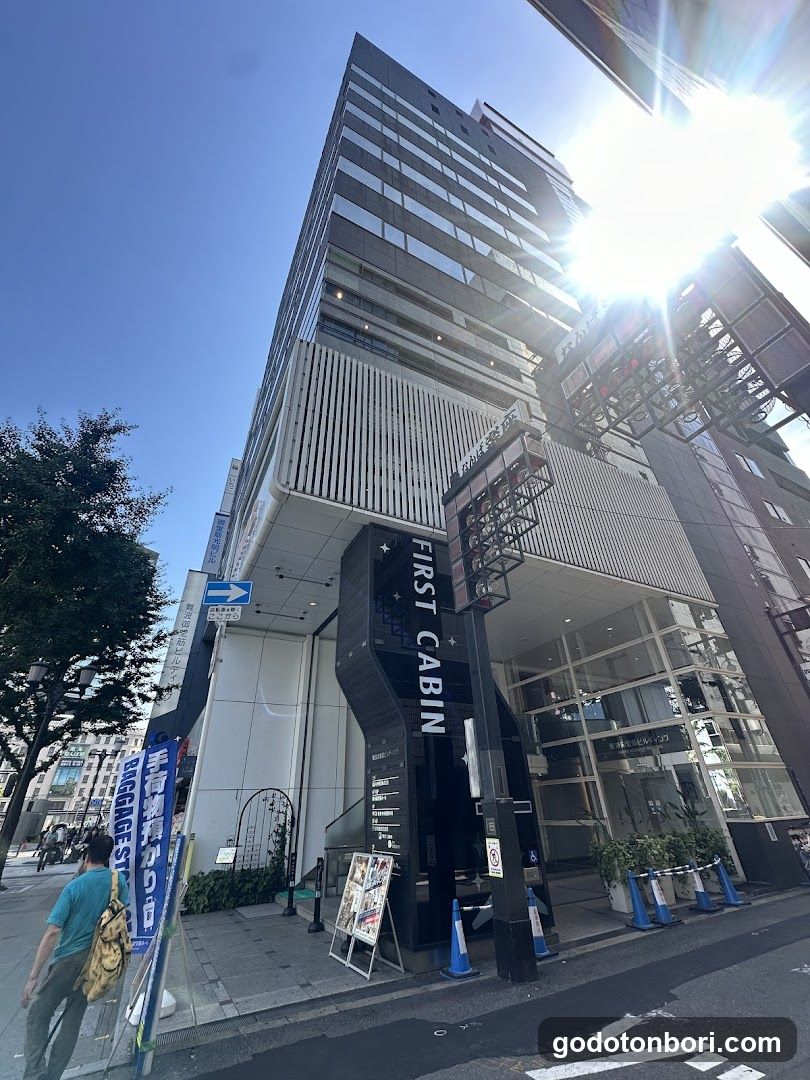 A modern building exterior with a prominent entrance labeled 'FIRST CABIN' in white vertical lettering on a black column. The facade features glass windows and metal grilles, with blue traffic cones placed near the sidewalk. A person walks by carrying a bag.