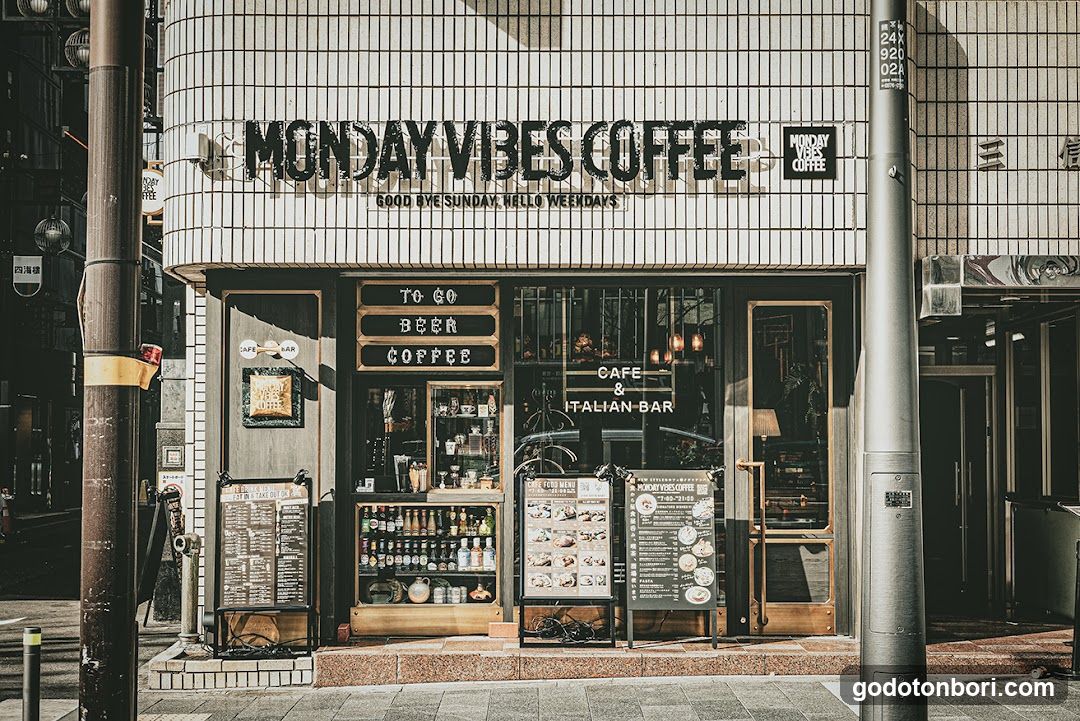A street-level view of a coffee shop with tiled facade and large glass windows displaying interior shelves. Signage reads 'MONDAY VIBES COFFEE' and 'GOOD BYE SUNDAY, HELLO WEEKDAYS'. Entrance flanked by menu boards.