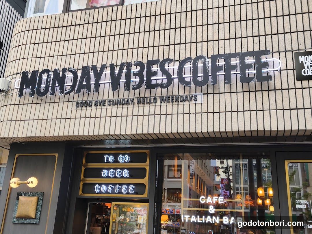 The image shows the exterior of a coffee shop named 'MONDAY VIBES COFFEE' with signage reading 'GOOD BYE SUNDAY, HELLO WEEKDAYS'. The storefront features large windows displaying interior elements and additional signs indicating it is also an Italian Bar.