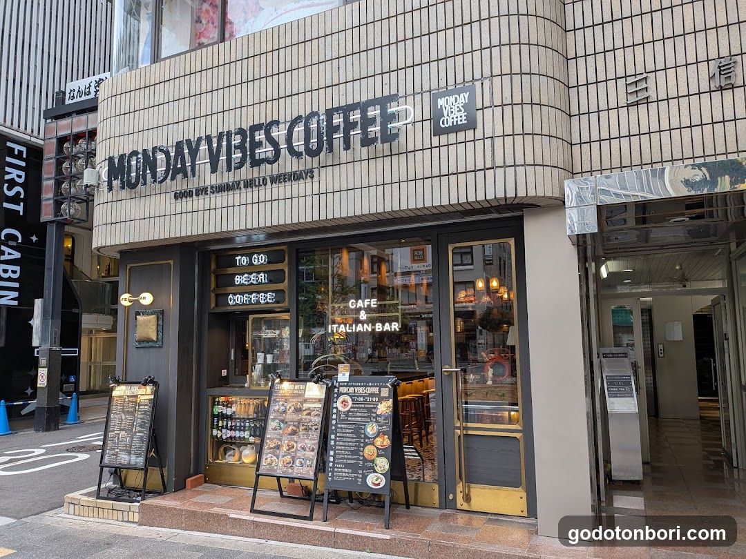 The exterior of Monday Vibes Coffee, a cafe and Italian bar located in an urban setting. The storefront features large glass windows displaying the interior ambiance with warm lighting and wooden furniture. Outside, there are several menu boards on stands showcasing various food items and prices. A digital signboard above the entrance displays operating hours.