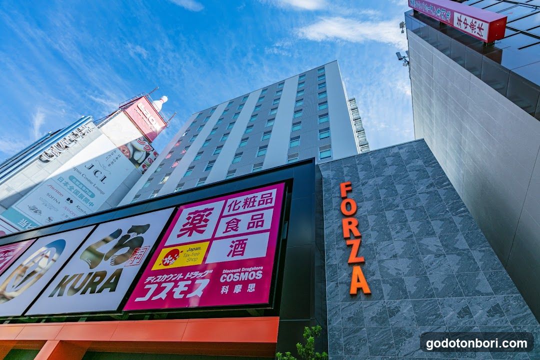 Low-angle shot of a city street featuring multiple buildings under a blue sky. Prominent signs include 'FORZA' in orange on a gray tiled wall, and large billboards advertising brands like KURA, Japan Cosmetics (COSMOS), and pharmacy items such as medicine, food, and alcohol. A red sign with Japanese characters is visible atop a glass building to the right.