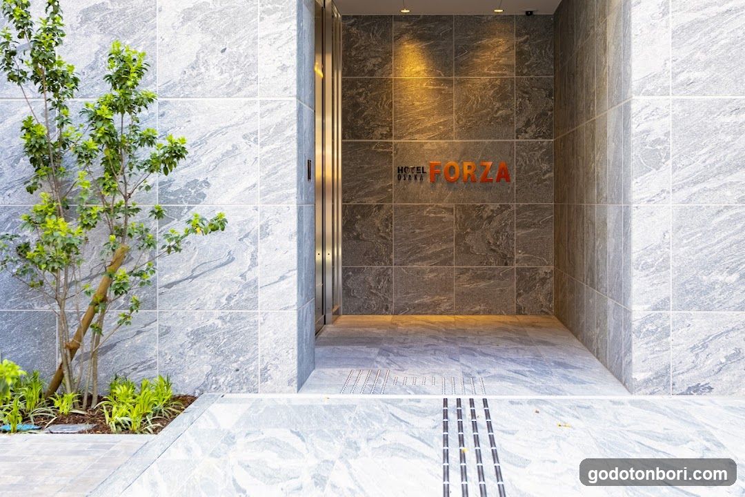 Exterior entrance of Hotel Forza featuring marble walls, a small tree on the left, and illuminated signage reading 'HOTEL FORZA' on the wall.
