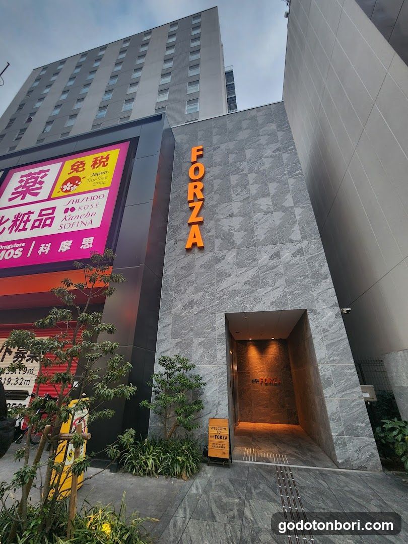 Photo of the physical exterior entrance and storefront facade of a building with 'FORZA' signage in orange letters on a gray tiled wall. A large pink signboard displays Japanese text including '薬', '免税', '化粧品', and 'Kanebo'. The entrance features a recessed doorway illuminated from within, flanked by small green plants and a yellow sign near the ground.