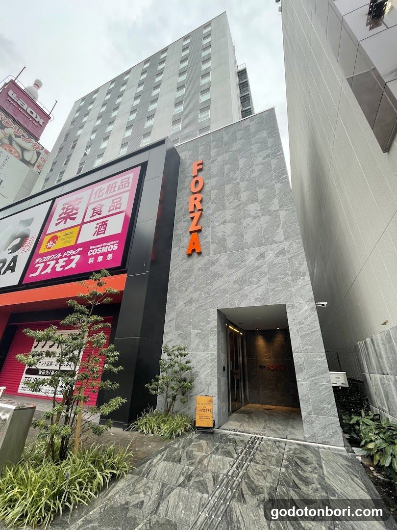 Photo of the physical exterior entrance and storefront facade of a building named FORZA, featuring gray stone cladding with vertical orange lettering. A large pink sign displays Japanese characters for cosmetics, food, medicine, and alcohol. Small trees and plants are planted along the sidewalk in front.