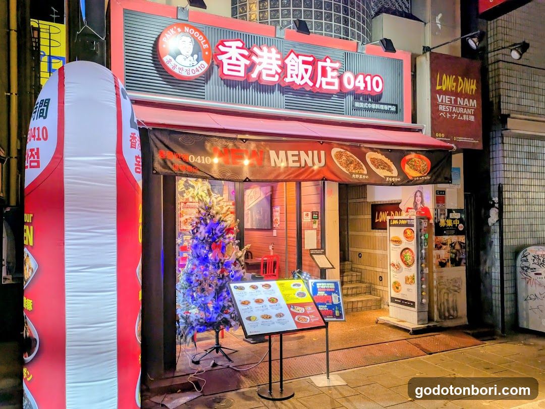 A brightly lit storefront at night featuring a red and white sign with Chinese characters '香港飯店' (Hong Kong Hotel) and the number '0410'. A large inflatable archway stands to the left, while a small decorated Christmas tree is placed near the entrance. The awning displays images of food items under 'NEW MENU', indicating a promotional display.