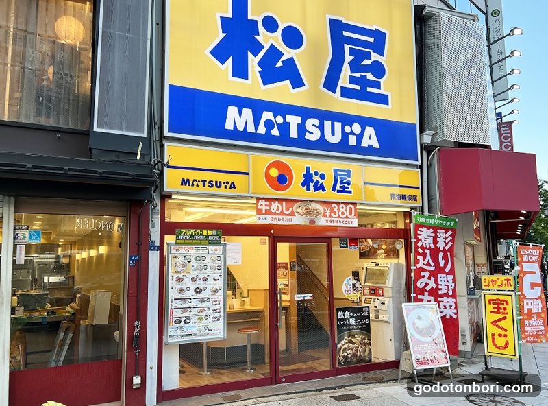 A brightly lit storefront with a large yellow and blue sign displaying '松屋' (MATSUYA) in Japanese characters and English letters. The entrance features glass doors leading into the interior, where warm lighting illuminates the space. A menu board is visible on the left side of the door frame, listing various food items. Additional signage includes promotional banners and advertisements for special dishes like '牛めし' (beef rice). The building facade has modern architectural elements, including metal grilles and illuminated signs.
