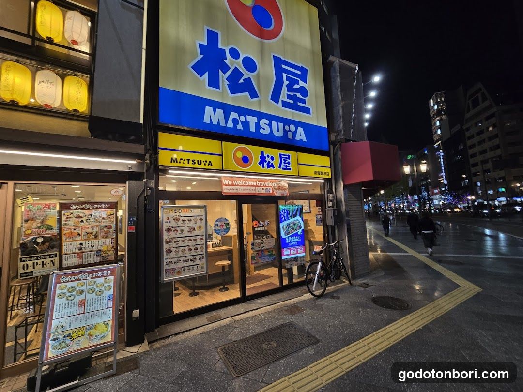 Night view of a Matsuya restaurant storefront with illuminated signage displaying Japanese characters and English text 'MATSUYA'. The entrance features glass doors, menu boards outside, and a bicycle parked nearby on the sidewalk.