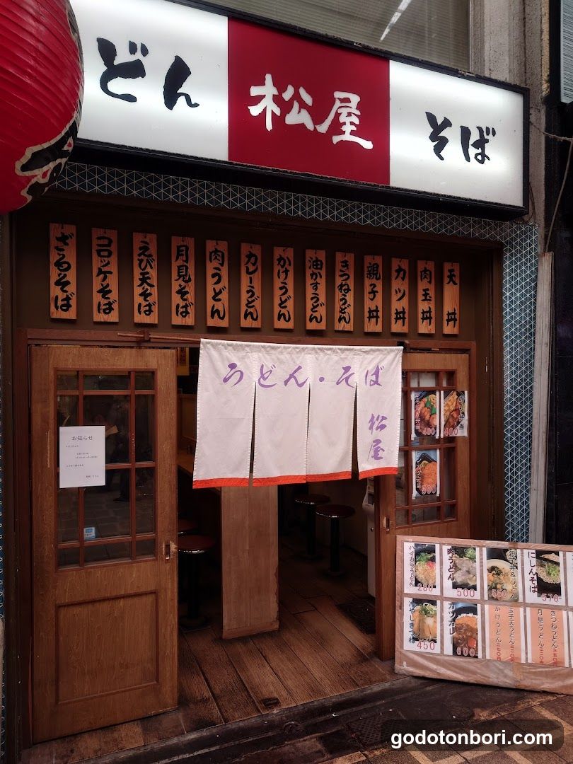 A traditional Japanese restaurant entrance with wooden doors and a hanging noren curtain displaying 'udon & soba'. Above, the sign reads 'don' in black on white, followed by red background with 'matsuya', and 'soba' in black. A vertical menu board lists various dishes like 'katsu soba', 'men men soba', 'chikuwara soba', 'nori soba', 'tama soba', 'shiro soba', 'yaki soba', 'kara soba', 'kara udon', 'kara udon', 'oyasoban', 'kushi soba', 'katsu soba', 'niku soba', and 'ten soba'. A small signboard outside advertises 'udon & soba' with prices listed for different items.
