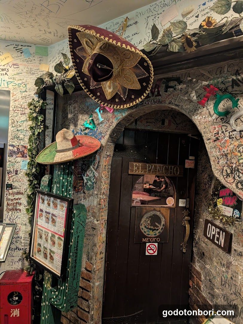 A rustic storefront entrance with a dark wooden door labeled 'EL PANCHO' and 'MEXICO'. The walls are covered in graffiti, stickers, and handwritten notes. Decorative elements include two sombreros (one large above the arch), green cactus-shaped pillars, and hanging sunflowers. A framed menu board is mounted on the left wall next to a red fire extinguisher box.