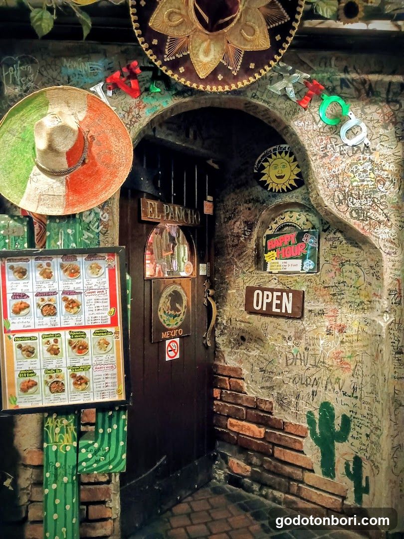 A rustic, graffiti-covered entrance to a Mexican restaurant featuring a dark wooden door with 'EL PARQUE' and 'MEXICO' signs above it. Large sombreros hang on the wall, and an 'OPEN' sign is visible next to a cactus mural. A menu board stands outside displaying various food items.