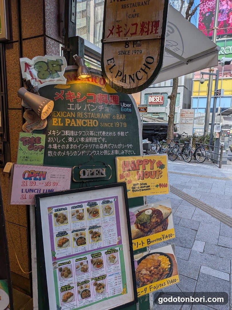 A street-level view of the exterior entrance to El Pancho, a Mexican restaurant and bar established in 1979. The facade features multiple signs including a hanging wooden sign with Japanese text 'メキシコ料理' (Mexican Cuisine) and English lettering. A large green chalkboard-style menu board lists hours, Uber Eats availability, and promotional offers like 'Happy Hour'. Below it is an open framed menu displaying burritos, fajitas, and other dishes with prices in yen. Additional signage indicates lunch times from 11:00-23:00.