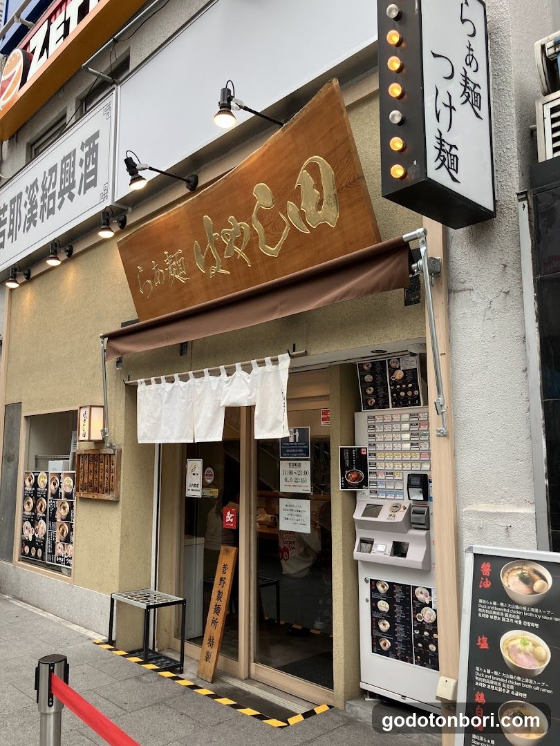 A shop front with a wooden sign reading 'ramen' and Japanese characters, white noren curtains at the entrance, and a menu board on the wall.
