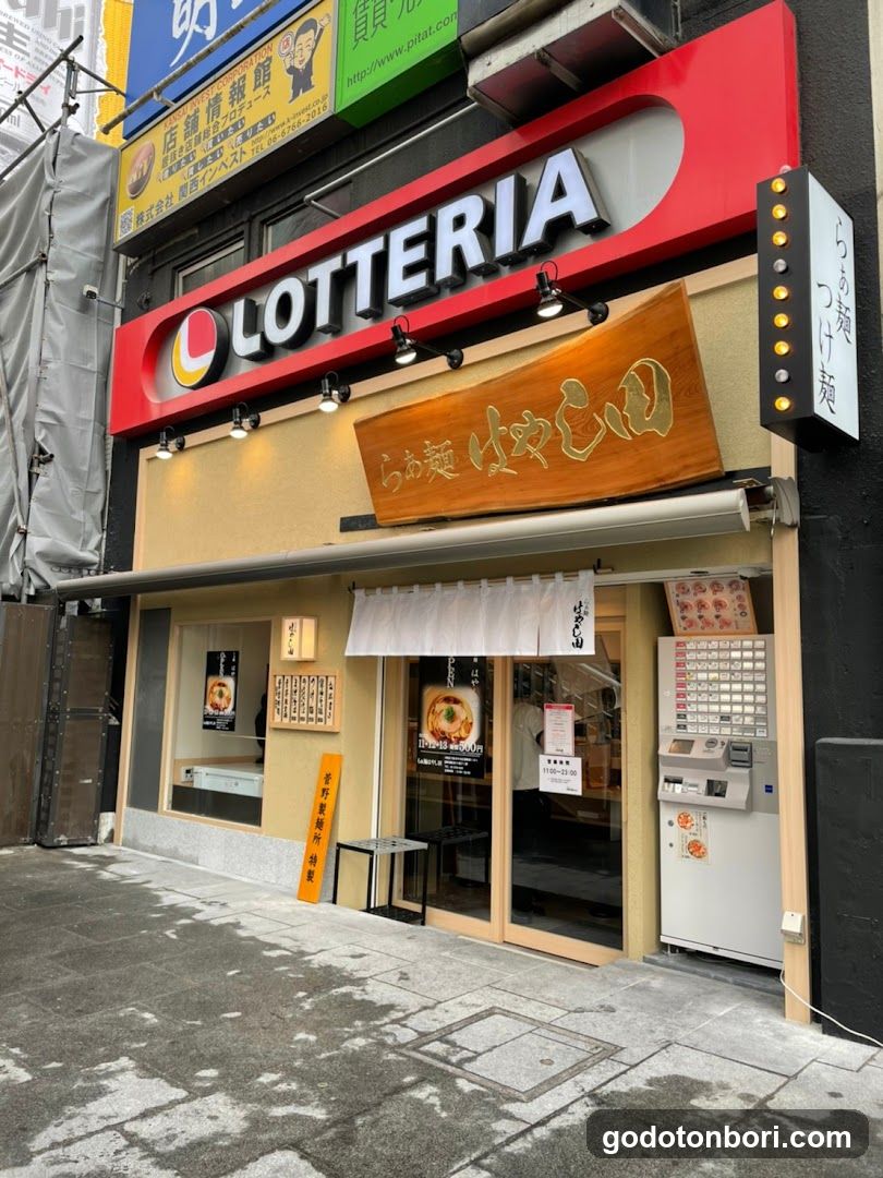 The image shows the exterior of a Lotteria ramen shop with prominent signage displaying 'LOTTERIA' and Japanese characters indicating it serves ramen. There is an ATM machine to the right, a small table near the entrance, and posters on the windows advertising food items.