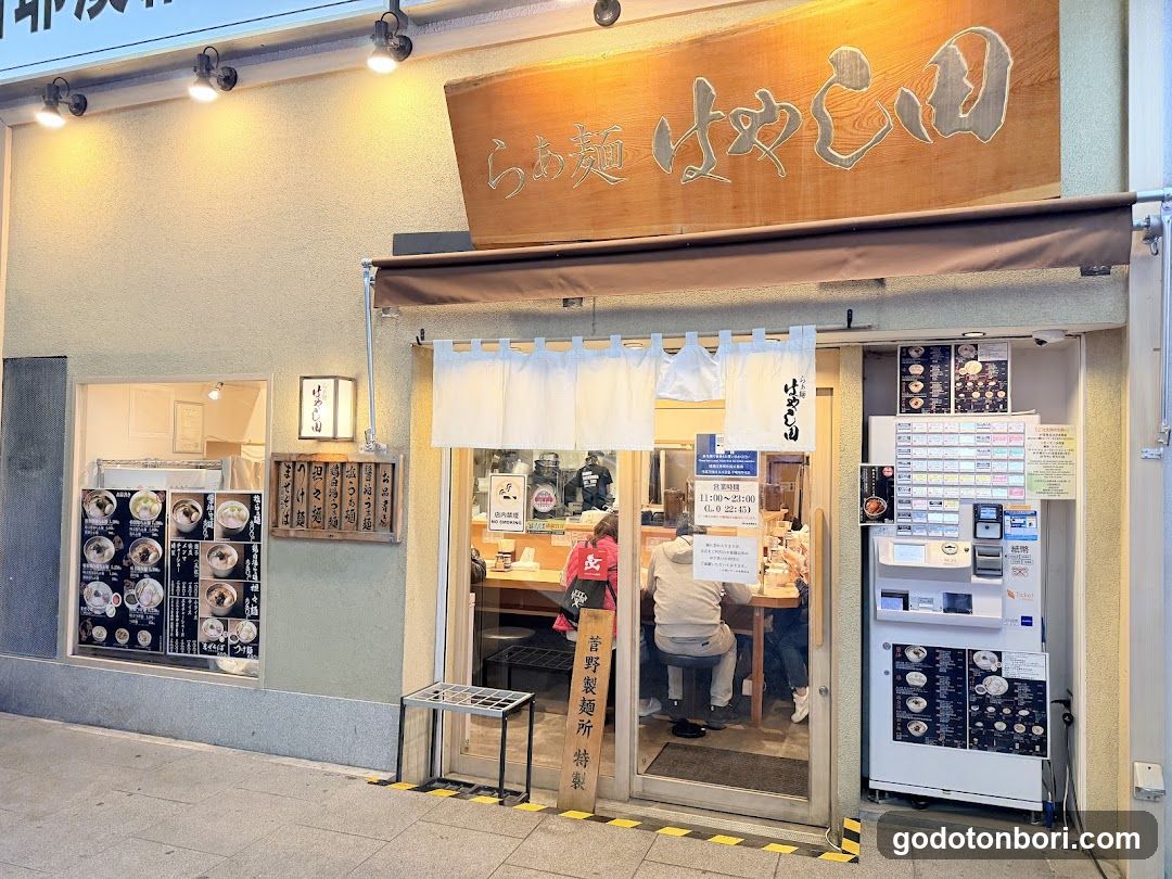 A small ramen shop with a wooden sign reading 'ramen hayakido' above the entrance. White noren curtains hang at the door, and a menu board is visible inside. A self-service payment machine stands to the right of the entrance.