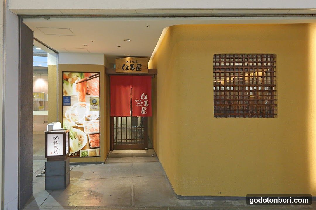 The image shows the entrance of a restaurant named 'Buta-ya' (buta meaning pig, suggesting pork specialty). A red noren curtain with white Japanese characters hangs at the doorway. Above it is a wooden sign reading 'Buta-ya'. To the left, there's a vertical illuminated signboard displaying the same name and logo. Adjacent to this is a large poster showcasing food items like sliced meat and bowls of soup or rice dishes. The wall on the right features a decorative square window made of wooden lattice panels. The interior appears warmly lit through the glass door.
