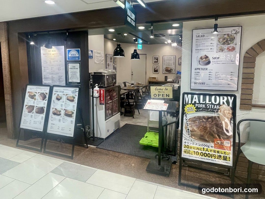 The image shows the entrance of a shop named 'Mallory Pork Steak'. There are several menu boards outside, including one listing pork steak options and another advertising a special offer with a large piece of meat. Inside, there is a counter area with a refrigerator displaying food items. The interior has tables and chairs set up for customers.