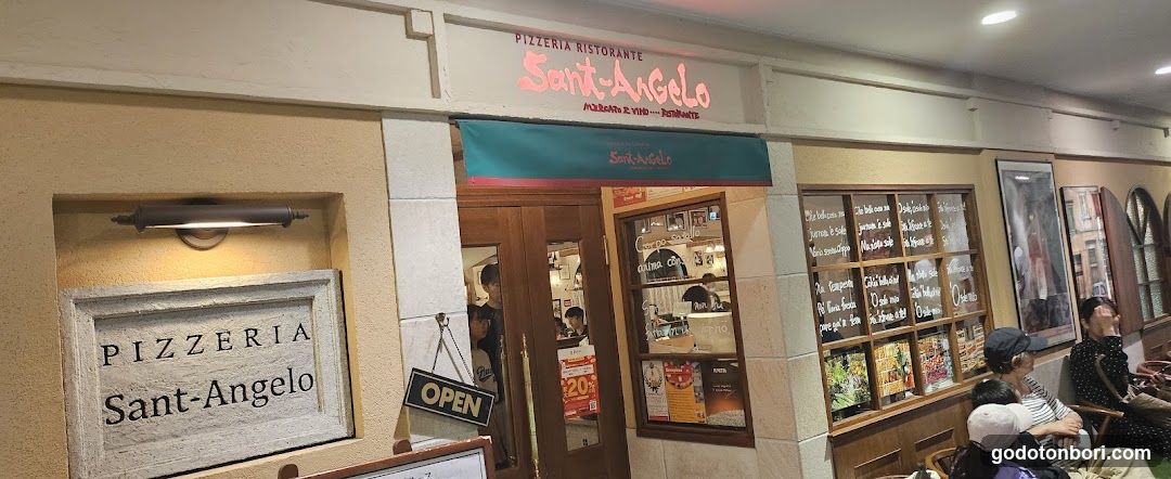 The image shows the exterior of Pizzeria Sant'Angolo, a restaurant with signage on the building facade. The entrance features glass doors and windows displaying menus and promotional posters. A chalkboard menu board is visible to the right of the entrance, listing various food items and prices. People are seated outside in chairs near the entrance.