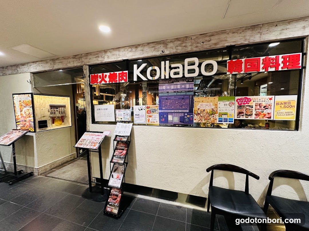 The image shows the storefront of a restaurant named 'KollaBo' with signage in both English and Japanese, indicating it serves Korean cuisine. The shop front features large glass windows displaying various posters and advertisements inside. There are menu boards outside the store, including one on a stand near the entrance. Two chairs are visible outside the shop.