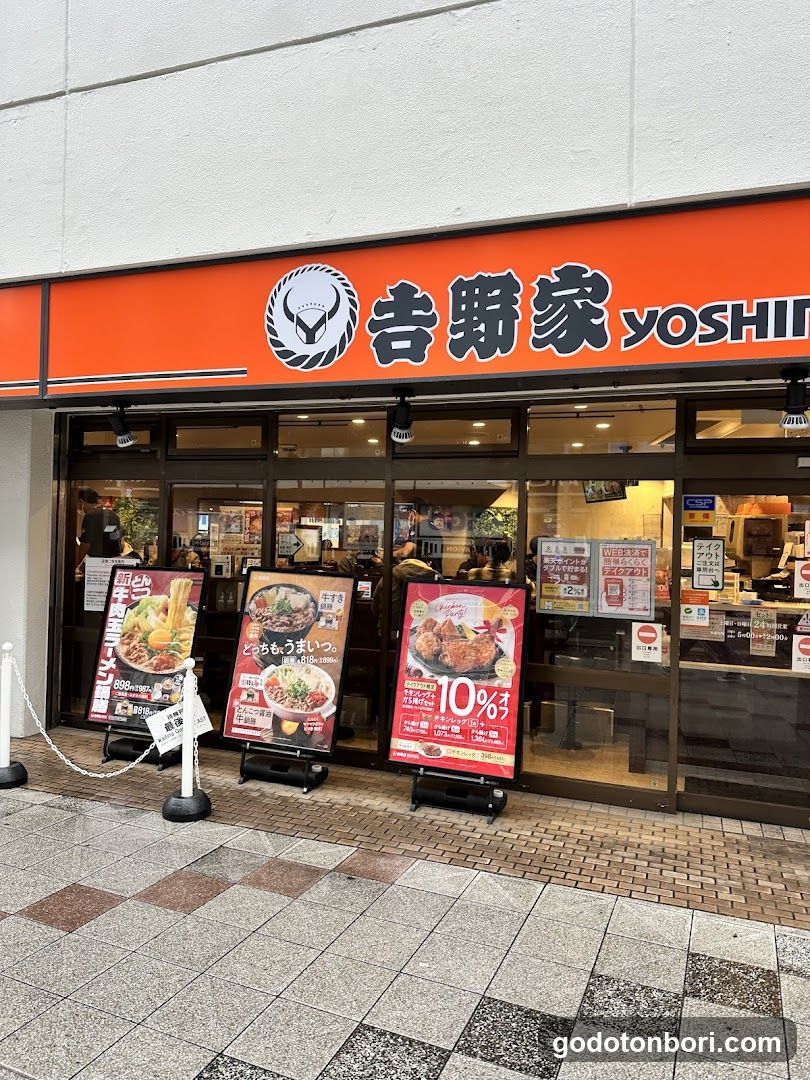 A street-level view of a Yoshinoya restaurant entrance, featuring an orange sign with the brand logo and name in Japanese characters (吉野家) and English (yoshinoya). Three A-frame promotional boards stand outside displaying menu items like ramen, tonkatsu, and other dishes with prices. The store interior is visible through glass doors, showing staff and customers inside.