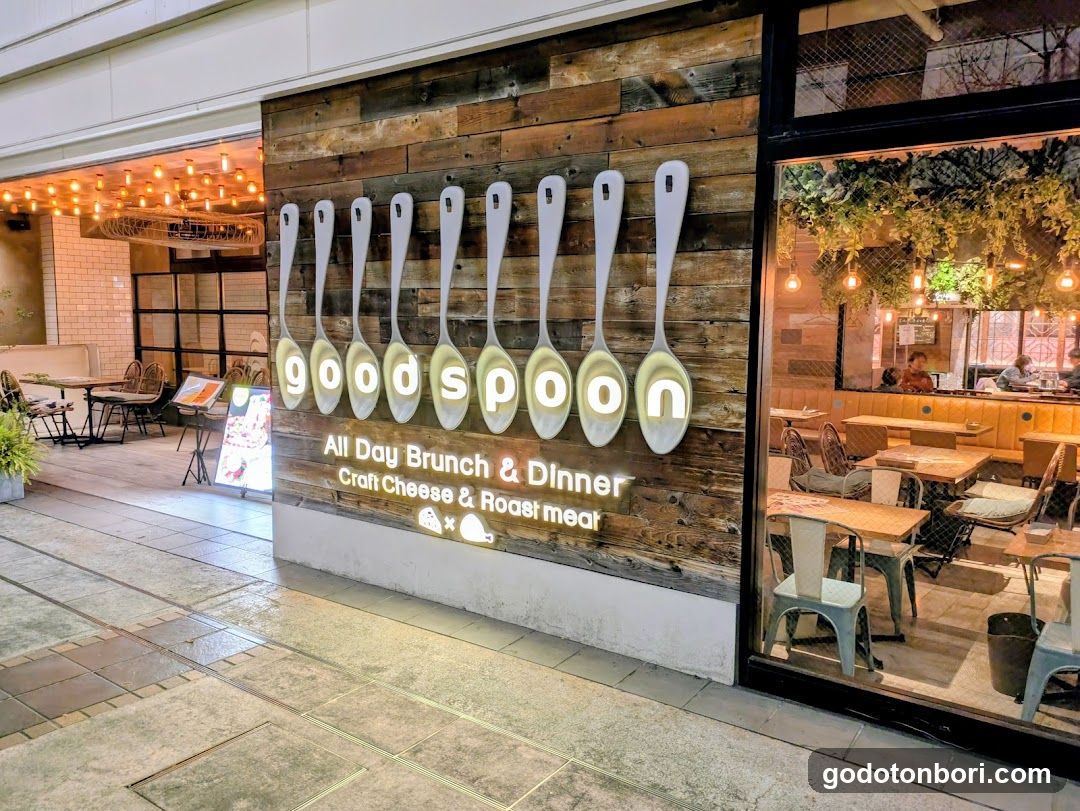 A storefront with wooden paneling and illuminated spoon-shaped signs spelling 'spoon'. The sign reads 'All Day Brunch & Dinner' and mentions 'Craft Cheese & Roast meat'. Inside, tables are set up under warm lighting with hanging greenery.