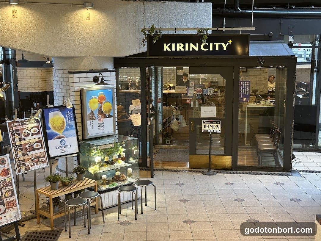 A modern storefront with glass doors and windows, displaying a 'KIRINCITY' sign above the entrance. Inside, staff members are visible behind counters. Outside, there are promotional posters featuring drinks and food items, along with small tables and stools arranged near the display cases.