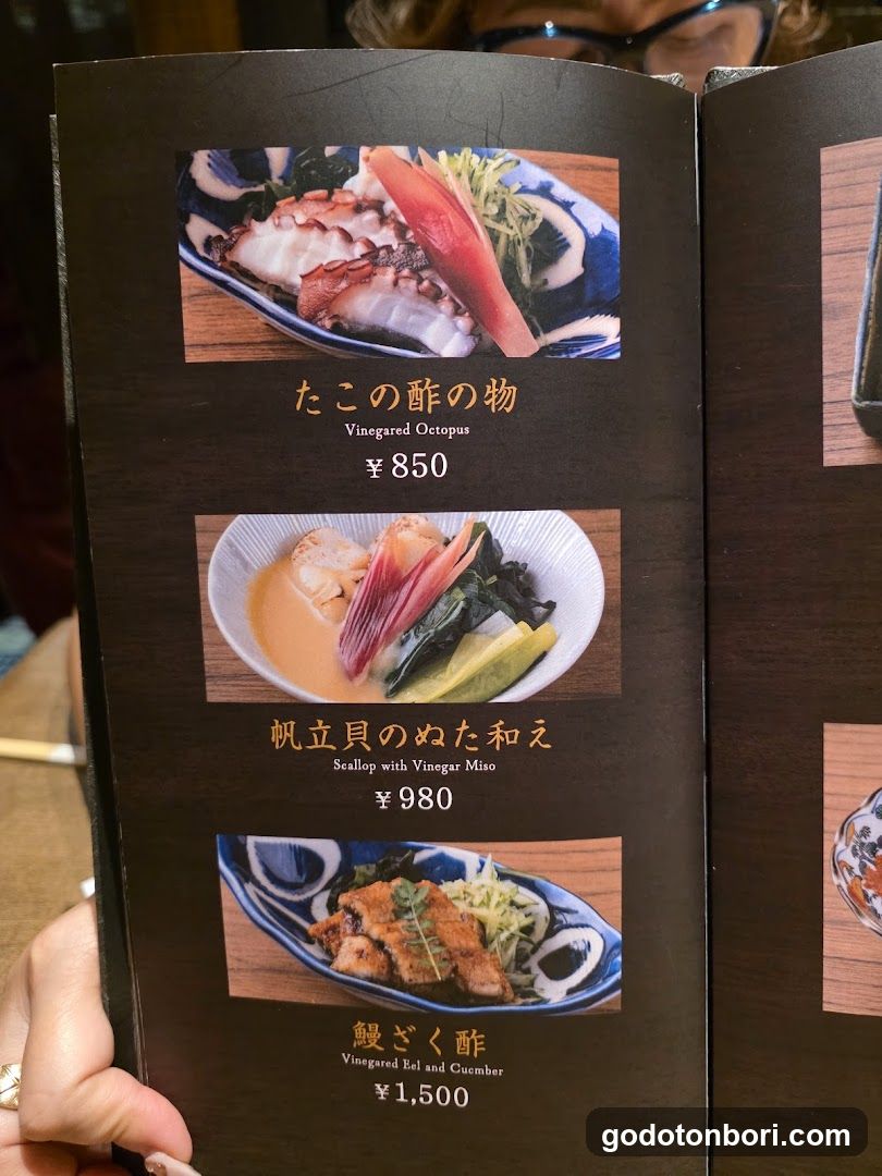 A close-up of a dark brown menu page held by a hand, displaying three appetizers with photos and prices in yen.