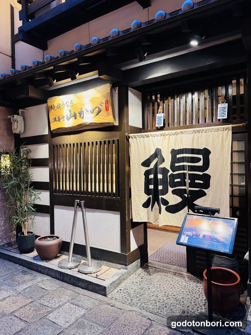 A traditional-style shop entrance with wooden slats and a large hanging banner displaying the kanji character '鰻' (eel). Above, another sign reads '地獄うなま 流香 山がつ'. A small potted plant sits to the left, while a digital menu board stands outside near the entrance.