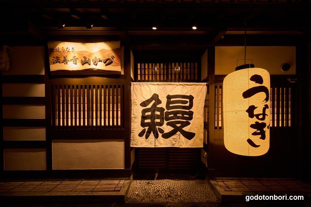 A warmly lit traditional Japanese shop entrance featuring wooden slatted doors, a hanging lantern with Japanese characters, and banners displaying large kanji characters. The scene suggests an authentic cultural or culinary establishment.