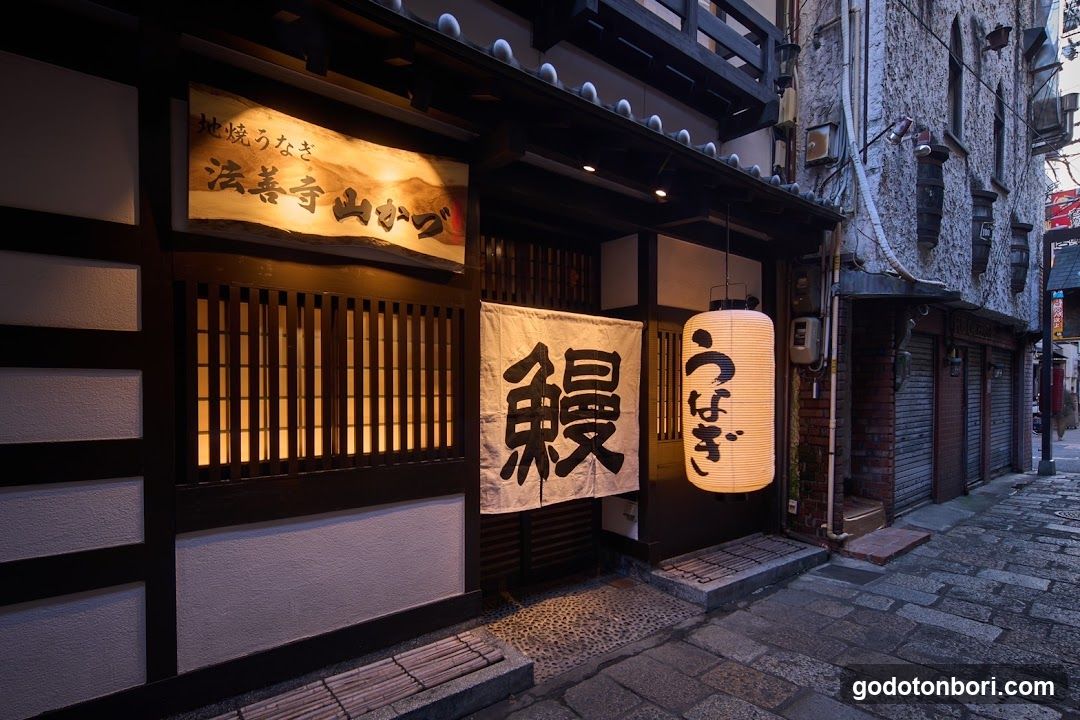 A traditional Japanese shop front with wooden architecture, featuring a lit lantern and hanging banners displaying Japanese characters. The entrance is narrow, flanked by stone pavement and adjacent buildings.