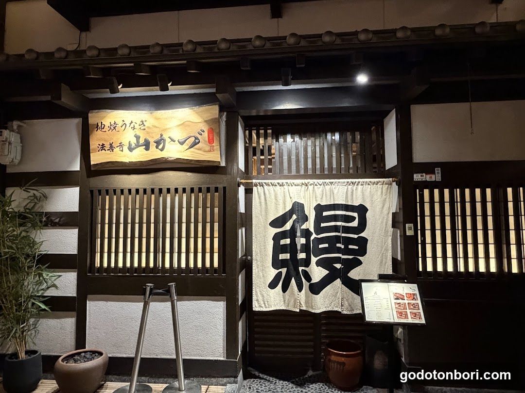A traditional Japanese shop front with wooden lattice doors and a hanging noren curtain displaying the kanji '鰻' (unagi). Above, a sign reads '地焼うなぎ 流善寺 山がぶ'. A small menu board is visible to the right of the entrance.
