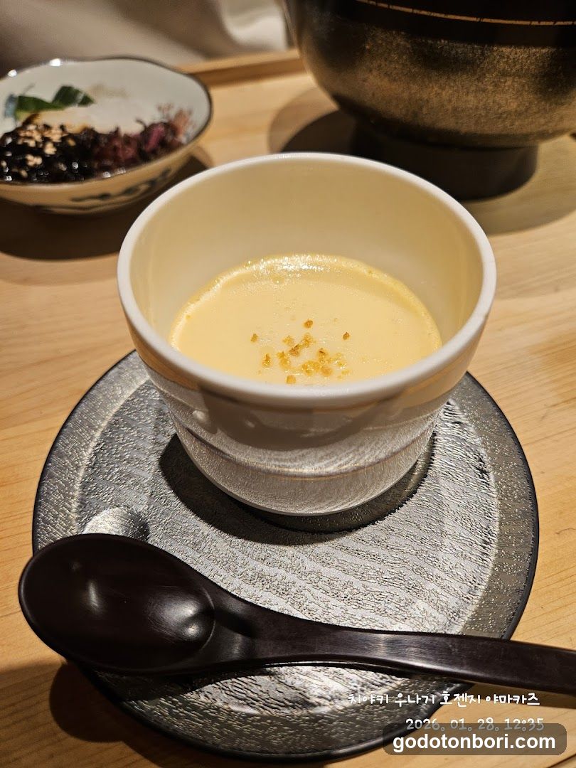 A close-up of a creamy yellow dessert in a white ceramic bowl, garnished with sesame seeds, placed on a textured gray plate with a dark wooden spoon beside it. In the background, there is a small dish containing black and purple toppings, possibly seaweed or fruit pieces, and part of a large metallic pot visible. The setting appears to be an indoor dining environment with warm lighting.