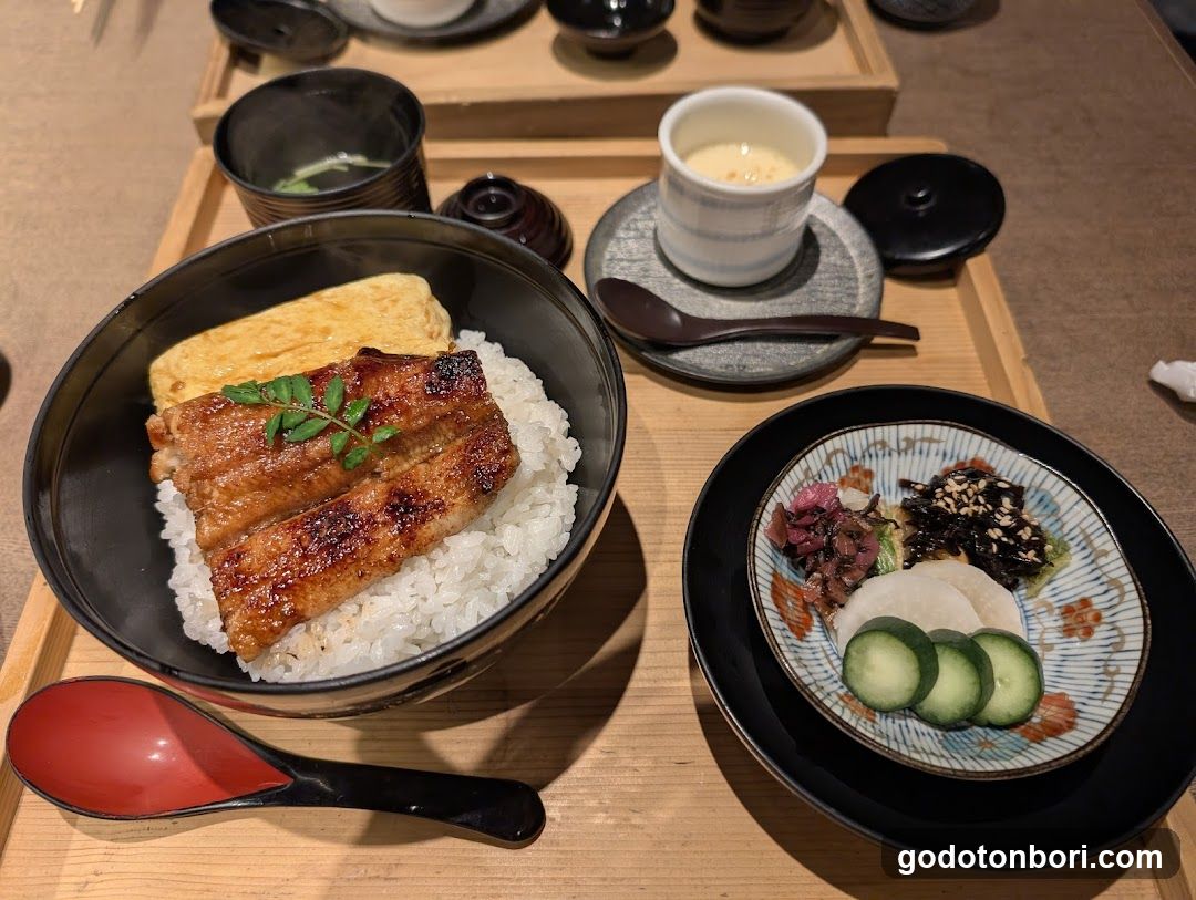 A close-up of a Japanese meal featuring grilled eel over rice in a black bowl, garnished with green leaves. Accompanying dishes include pickled vegetables, sliced cucumbers, and a small cup of soup or broth. A red-handled spoon rests beside the main dish, while another utensil is placed near a small cup on a wooden tray.