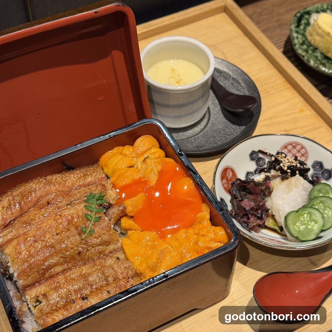 A close-up of a Japanese bento box featuring grilled eel (unagi) over rice, topped with bright orange unagi sauce and garnished with green herbs. Accompanying the main dish is a small bowl containing pickled vegetables including cucumber slices, seaweed salad, and possibly radish. A cup of light-colored soup or drink sits nearby on a dark saucer, along with a red spoon resting beside it. The setting appears to be indoors at a restaurant table.