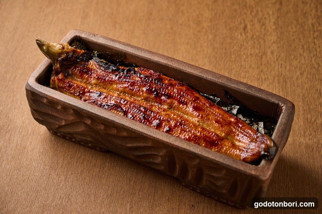 A single piece of grilled eel (unagi) served in a rectangular brown ceramic dish with textured sides, resting on dark seaweed or nori. The fish has a glossy, caramelized glaze with char marks along the skin. The dish sits on a light wooden surface under warm lighting.