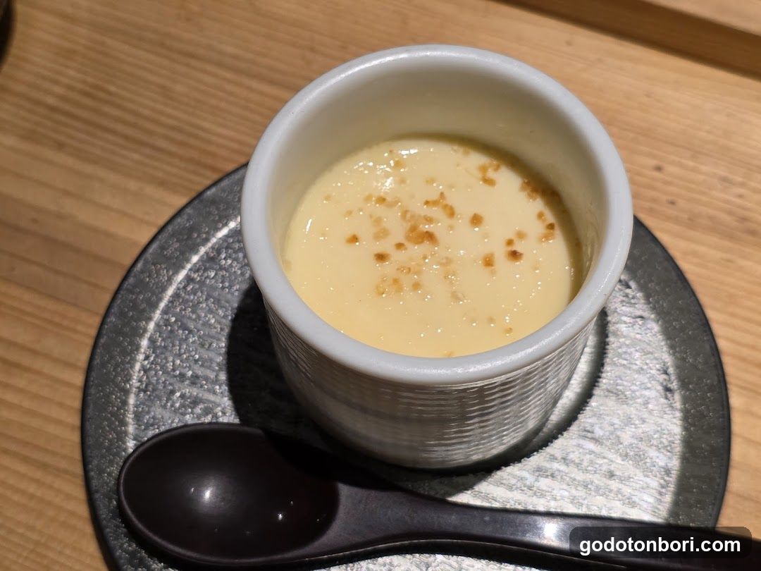 A close-up of a creamy egg custard dessert in a small white ceramic cup, garnished with brown crumbs, placed on a dark textured plate with a black spoon beside it.