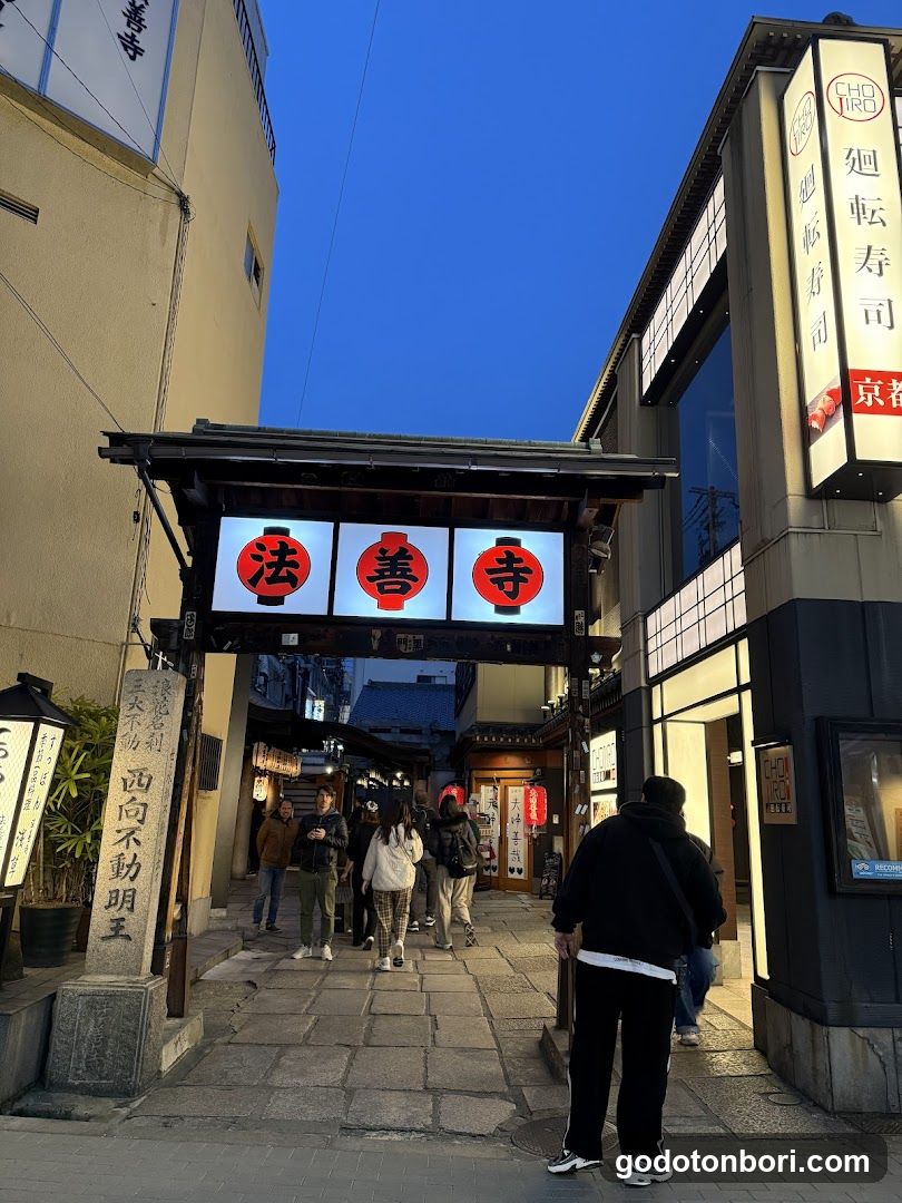 A street-level view of the entrance gate (torii) to Hossan-ji temple, featuring illuminated signs with Japanese characters '法善寺' (Hossan-ji). People are walking through the gateway under a clear blue sky. A shop front on the right displays vertical signage reading '廻転寿司' (Rotating Sushi), and another sign mentions '京都' (Kyoto). The scene captures traditional architecture mixed with modern commercial elements.