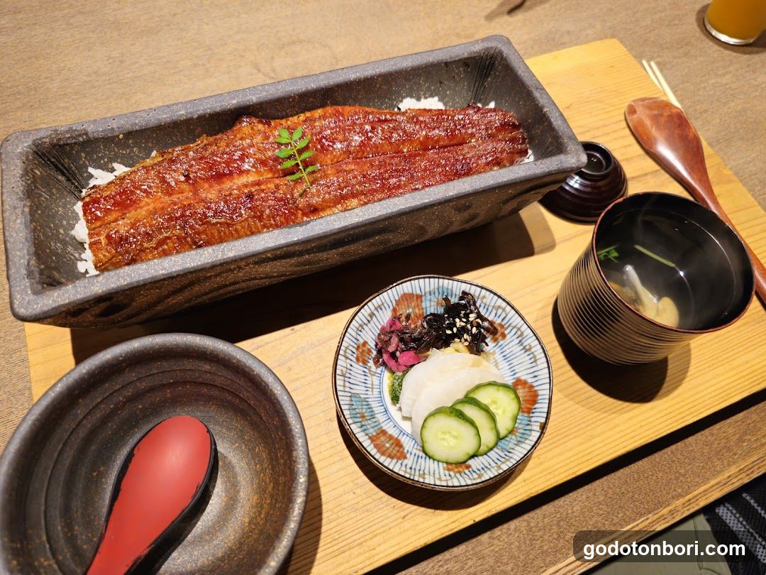 A close-up of a Japanese eel rice bowl (unagi don) served in a rectangular ceramic dish, garnished with green herbs. Accompanying items include a small plate with pickled vegetables and cucumber slices, a wooden spoon, a dark bowl with chopsticks, and a cup of tea or broth on a light wooden tray.
