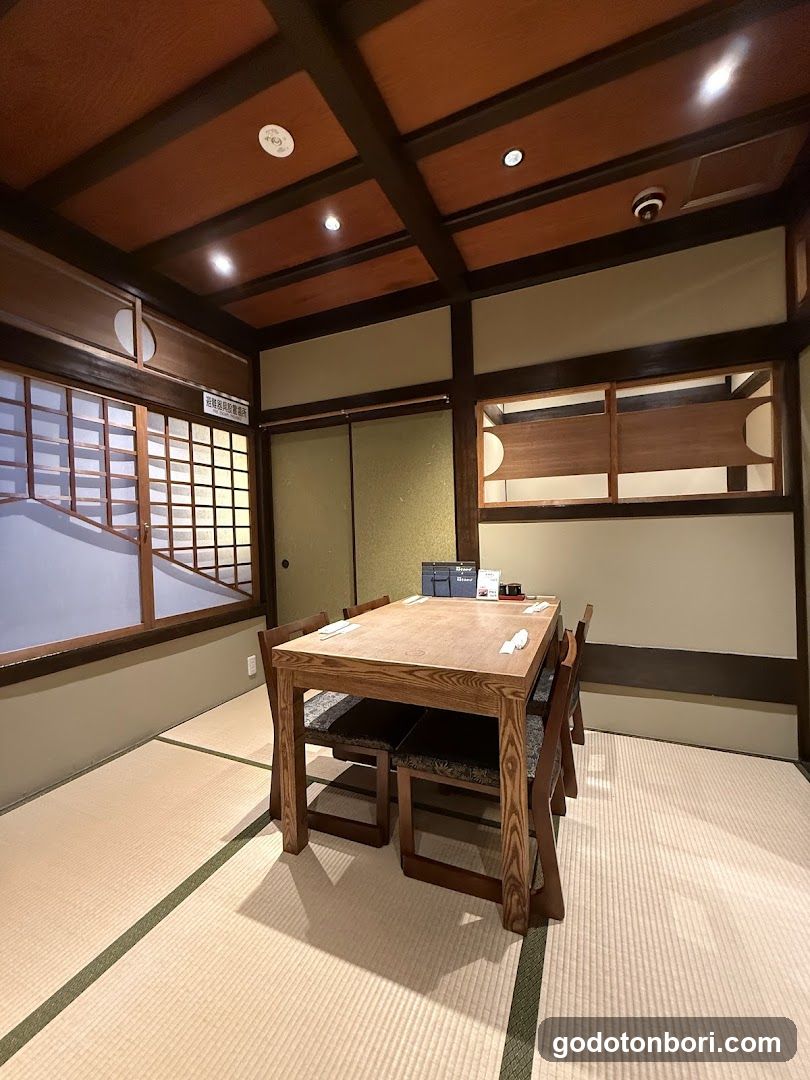 A traditional Japanese room with tatami flooring, wooden beams, and shoji screens. A low wooden table is set with napkins and small items, surrounded by matching chairs. The walls feature sliding doors and a window with grid panes.