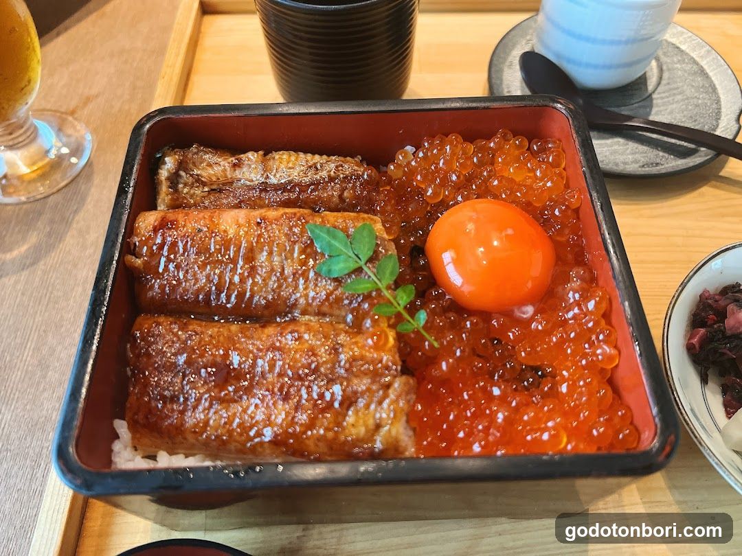 A close-up of a Japanese rice bowl featuring grilled eel (unagi) slices, bright orange salmon roe (masago), and a raw egg yolk on top. A sprig of green herb garnishes the dish. The bowl sits on a wooden table with other dishes in the background.