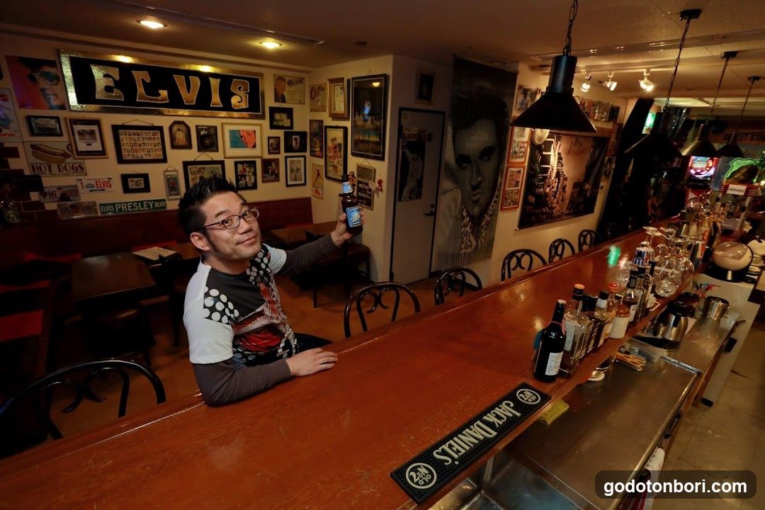 A man sits at a wooden bar in an Elvis-themed establishment, holding a beer bottle. The walls are decorated with framed photos, posters of Elvis Presley, and signs including 'ELVIS' and 'HOT DOGS'. A Jack Daniel's branded mat is visible on the bar counter.