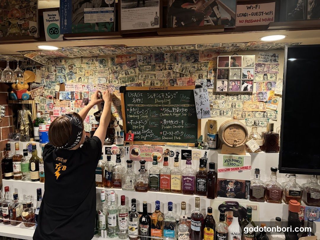 A bar interior featuring a wall covered in numerous banknotes, framed photos, and posters. A staff member wearing headphones is reaching up to adjust items on the wall. Below, shelves are stocked with various liquor bottles. A chalkboard menu lists drinks like Hamburg Steak and Chicken & Veggie Soup.