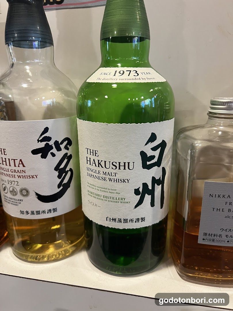 A close-up view shows three bottles of Japanese whisky lined up on a shelf. The central bottle is Hakushu Single Malt, with labels indicating it was established in 1973. To the left is a bottle labeled 'Shita', and to the right is another bottle partially visible with 'Nikka' branding.
