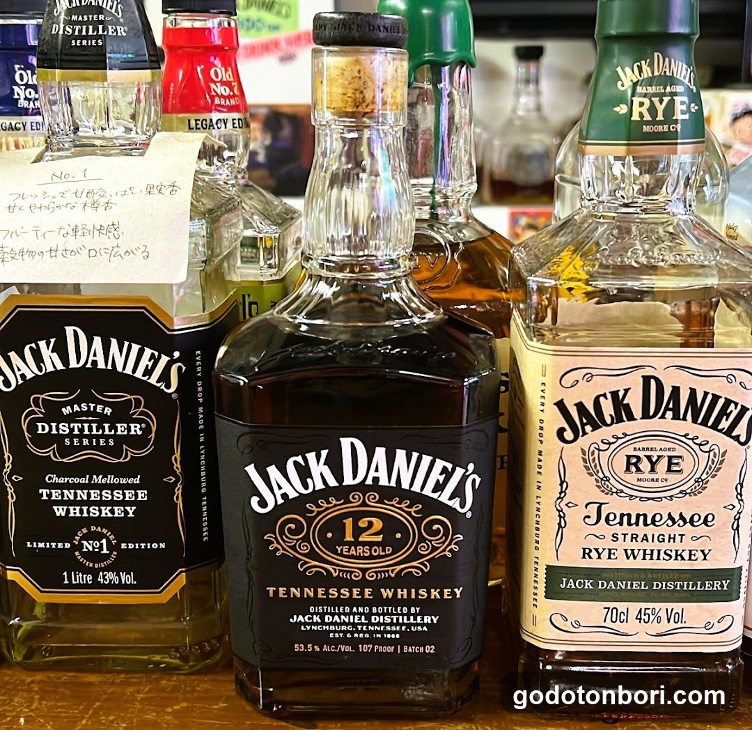 Close-up of several Jack Daniel's whiskey bottles on a wooden surface, including Master Distiller Series Charcoal Mellowed Tennessee Whiskey No.1, 12 Years Old Tennessee Whiskey, and Barrel Aged Rye Straight Rye Whiskey. A handwritten note in Japanese is attached to one bottle.