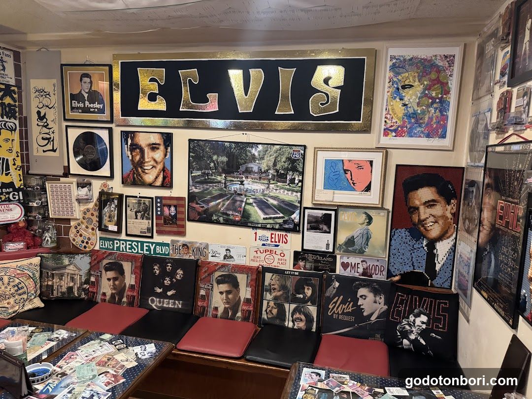 Interior shot of a room filled with Elvis Presley memorabilia, including framed posters, vinyl records, and plush cushions featuring his image. A large 'ELVIS' sign hangs above the wall, which is adorned with various photos and artwork related to the singer.