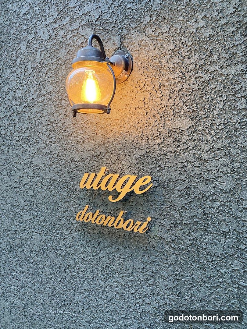 A close-up photo of a textured gray stucco wall featuring an illuminated vintage-style lantern light fixture mounted above two gold-colored cursive signs reading 'utage' and 'dotonbori'. The lighting casts a warm glow on the rough surface, suggesting evening or low-light conditions. No menu or interior details are visible.