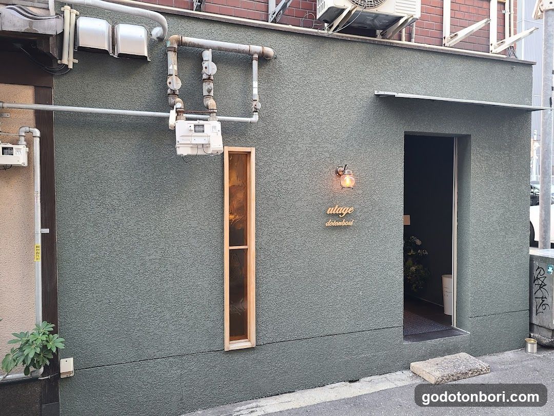 Green textured facade with vertical wooden window, entrance door, and 'alage' sign above door.